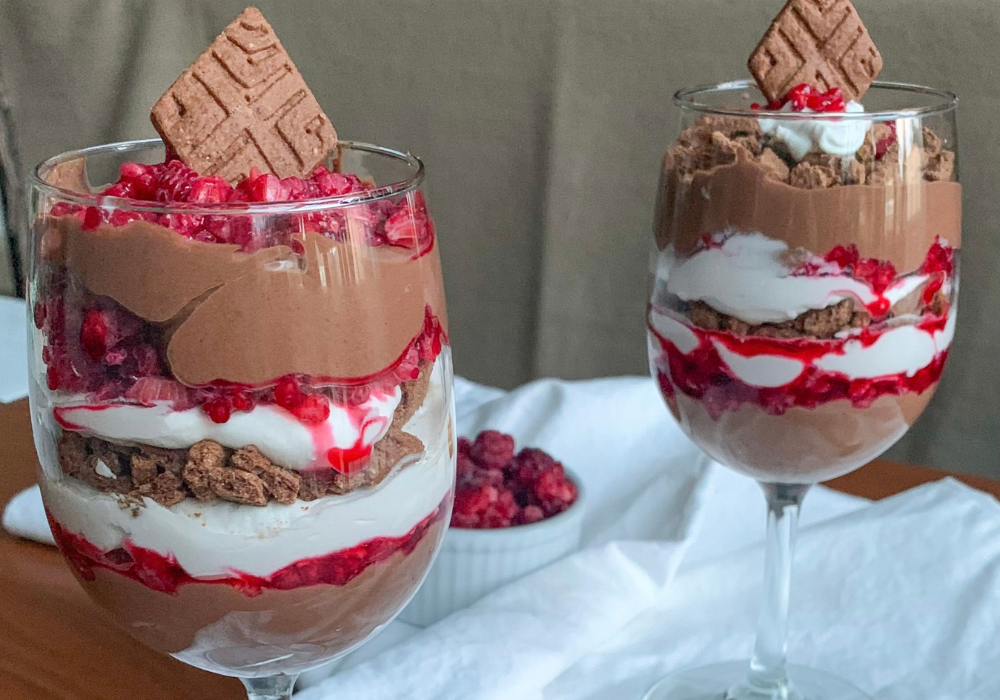 This image shows the ultimate dessert for a plant-based Valentine's dinner: a close up of chocolate raspberry parfaits with coconut whipped cream and chocolate cookie crumbs in two wine glasses.