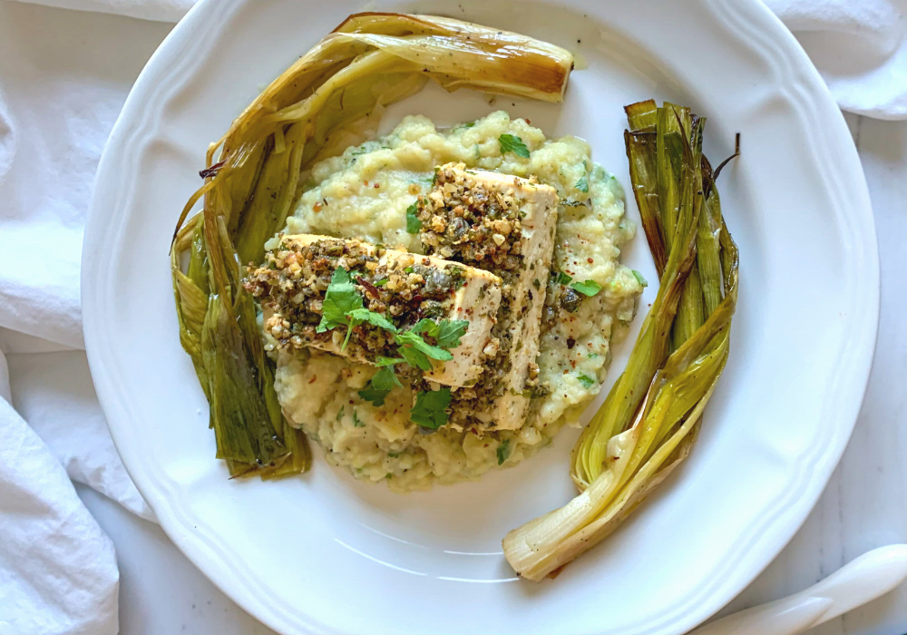 This image shows the perfect main course for a plant-based Valentine's dinner: lemon caper herb-crusted tofu on top of herbed mashed potatoes, with olive oil roasted leeks, and shallot butter sauce on a plate.