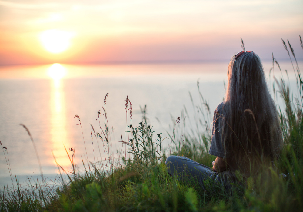 This image shows a woman peacefully sitting in nature watching the sunrise. Getting out in nature is a great stress management technique.