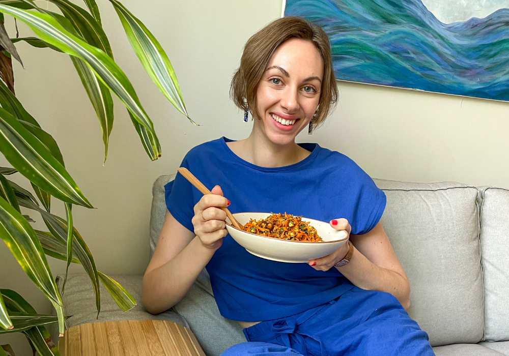 This image shows Fiona sitting on a couch holding a bowl of salad and smiling.