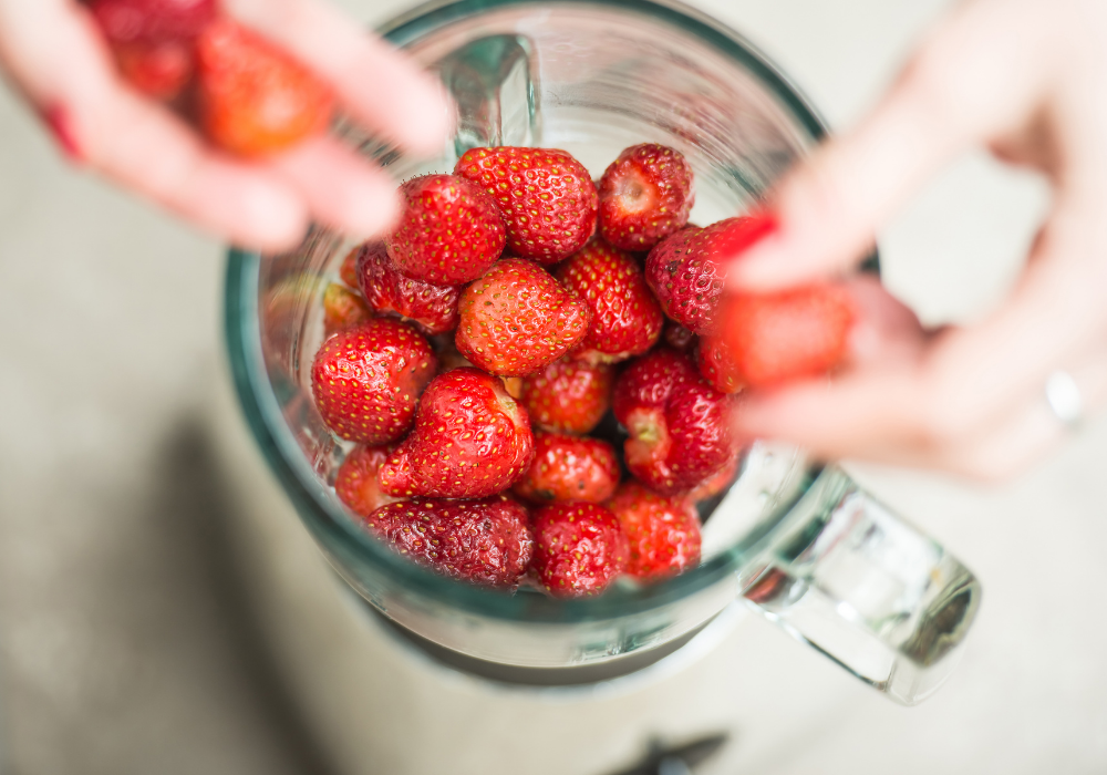 This image shows a powerful blender with hands putting strawberries into it to be blended.