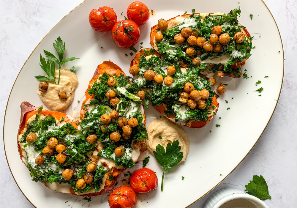 This image shows a delicious plant-based meal that features stuffed sweet potatoes loaded with protein-rich hummus, sauteed greens, and crispy chickpeas. Roasted cherry tomatoes sit on the side.