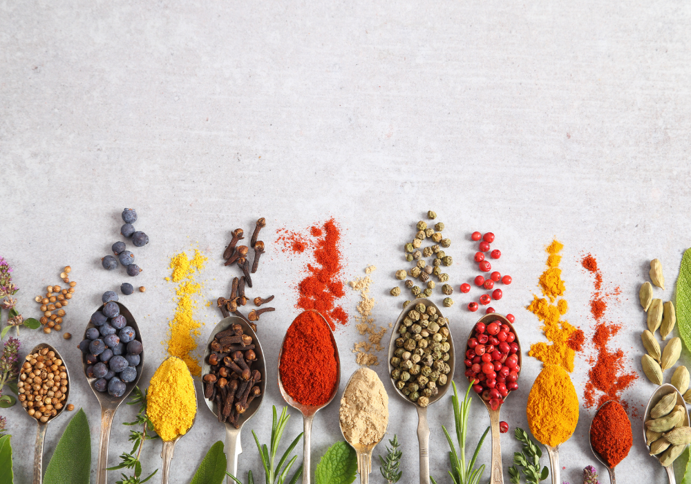 Image of a row of different spices coming off of spoons on a light grey surface.