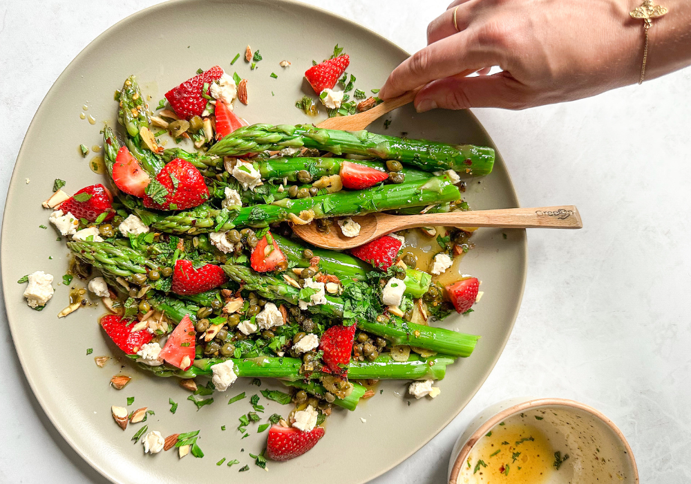 This image shows a strawberry asparagus salad on a platter with a hand holding a serving spoon.
