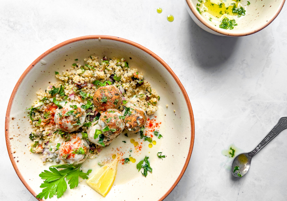 This is an image of a white bowl with quinoa, plant-based meatballs, and a creamy white sauce with green herbs scattered over top.