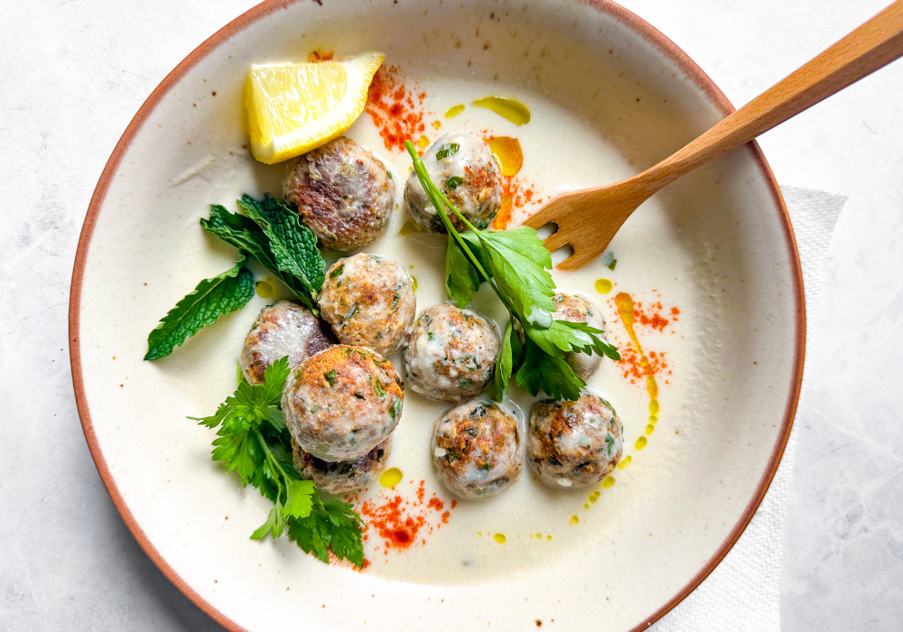 This is an image of a white bowl with meatballs in a creamy white sauce with green herbs scattered over top.