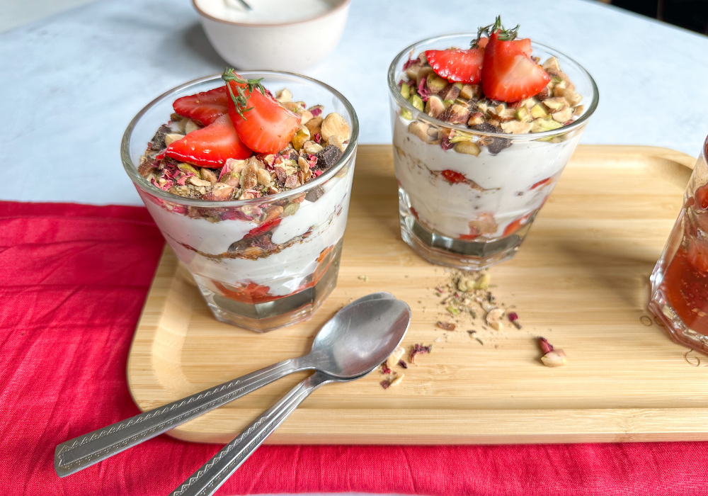 This image shows a side-view of two glasses of strawberry cream parfait, the third course of this seasonal spring menu. The glasses sit on a wooden tray on top of a bright pink cloth against a white background. 