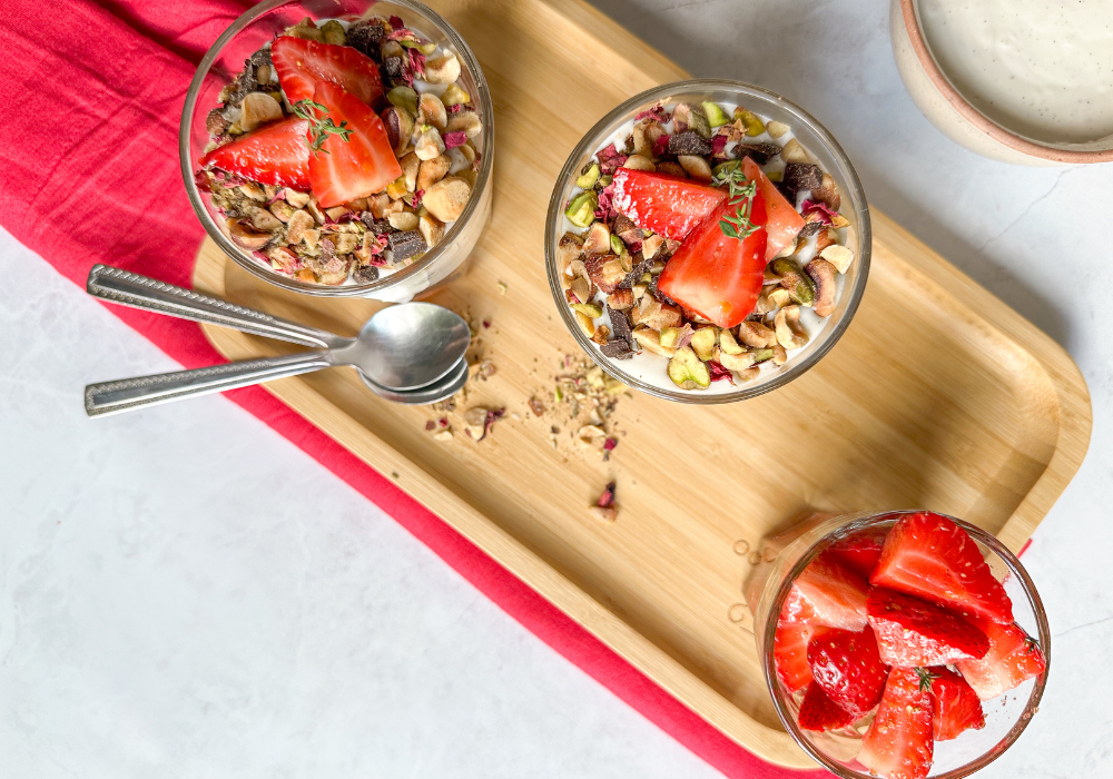 This is an overhead image of two glasses of of strawberry & cream parfaits. The parfaits sit on a wood tray with a bright pink cloth underneath on a white background.