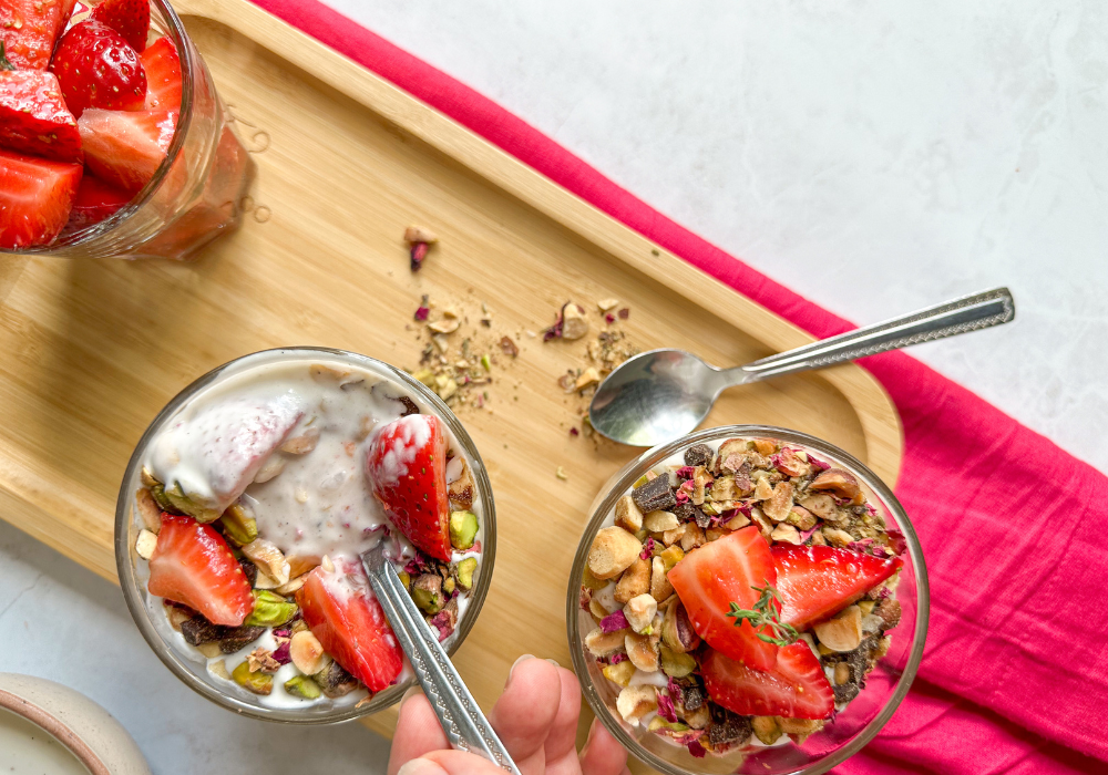 This is an overhead image of two glasses of of strawberry & cream parfaits with a hand scooping up some of the cream. The parfaits sit on a wood tray with a bright pink cloth underneath on a white background.