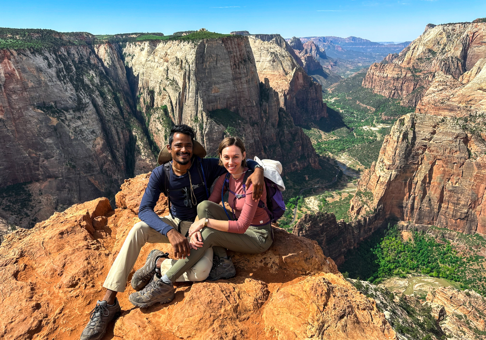 Fiona and her husband, Raju, sitting at the edge of a cliff with Zion Canyon National Park in the background.