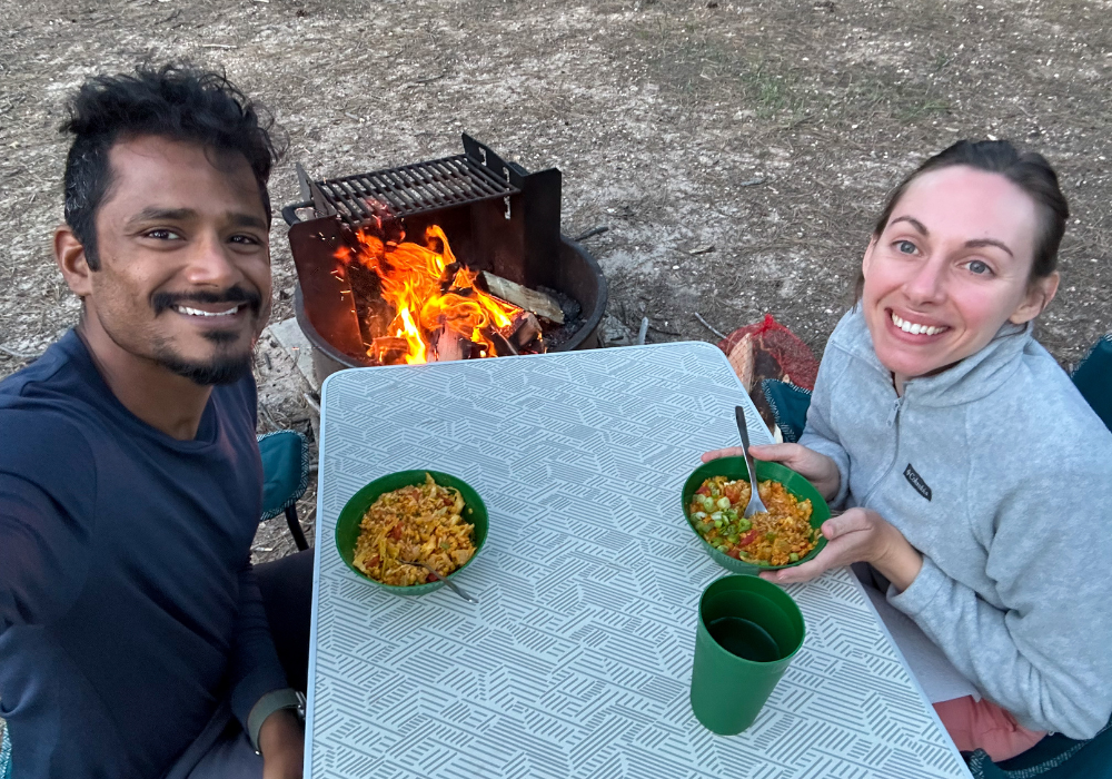 A mixed race couple enjoying bowls of red lentil quinoa stew next to a campfire.
