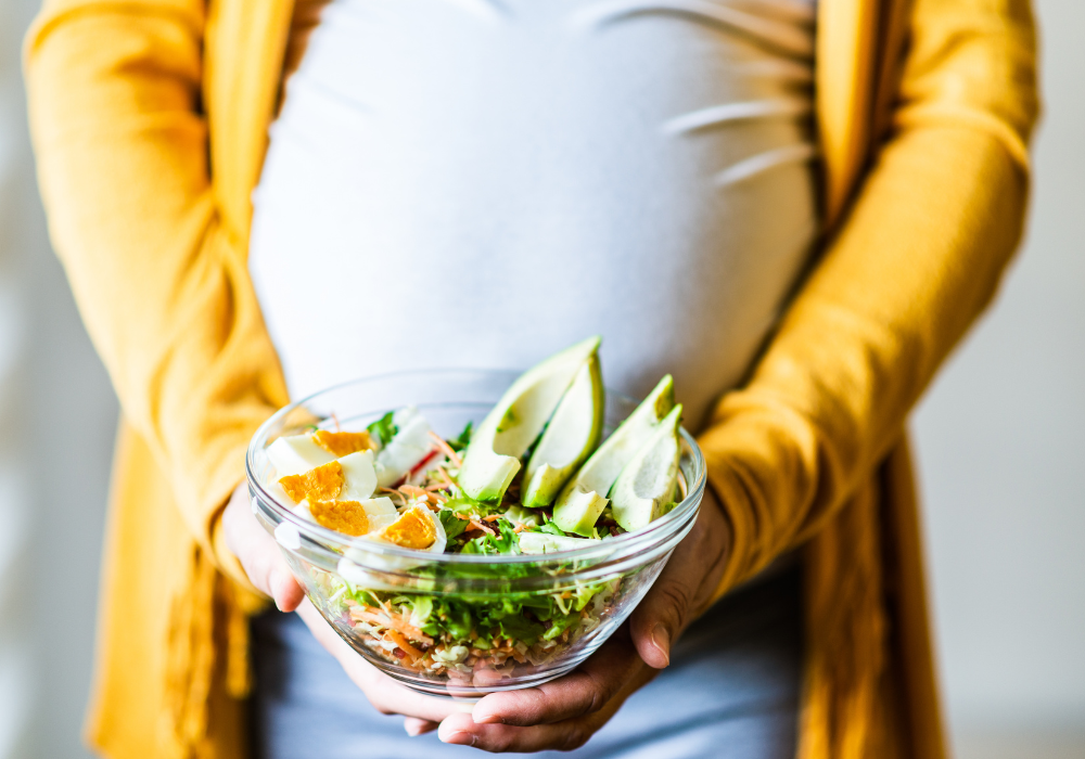 This image shows a pregnant woman in a yellow sweater holding a bowl of real foods that promote optimal nutrition during pregnancy. 